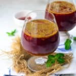 A close-up of two glasses filled with dark red Gajar Kanji, topped with spices, placed on a burlap mat with mint leaves beside them. A small bowl and more mint leaves are in the background.