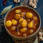 A bowl of spicy Kashmiri Dum Aloo with baby potatoes in a rich, red gravy, surrounded by whole spices like bay leaves, dried red chilies, ginger, and fennel seeds on a dark background.