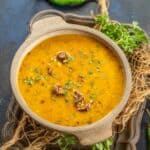 A bowl of urad dal garnished with herbs and dried red chilies, placed on a rustic tray with coriander leaves, a green chili, and a lemon wedge nearby.