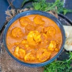 A black bowl filled with Goan Prawn Curry, sits on a rustic brown mat, garnished with fresh herbs, with naan bread and green chilies in the background.