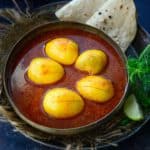 A bowl of spicy Kolhapuri Egg Curry with four boiled eggs in rich red gravy, served with two folded pieces of flatbread on the side, garnished with herbs and a lime wedge.