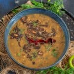 A bowl of Panchmel Dal topped with fried onions and a dried red chili, garnished with fresh herbs, sits on a rustic tray with jute fabric, fresh green chilies, and coriander leaves nearby.