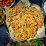 A basket lined with paper holds several pieces of crispy, golden-brown Sindhi Koki, garnished with herbs. Sides of chutneys and yogurt are placed nearby on a dark table.