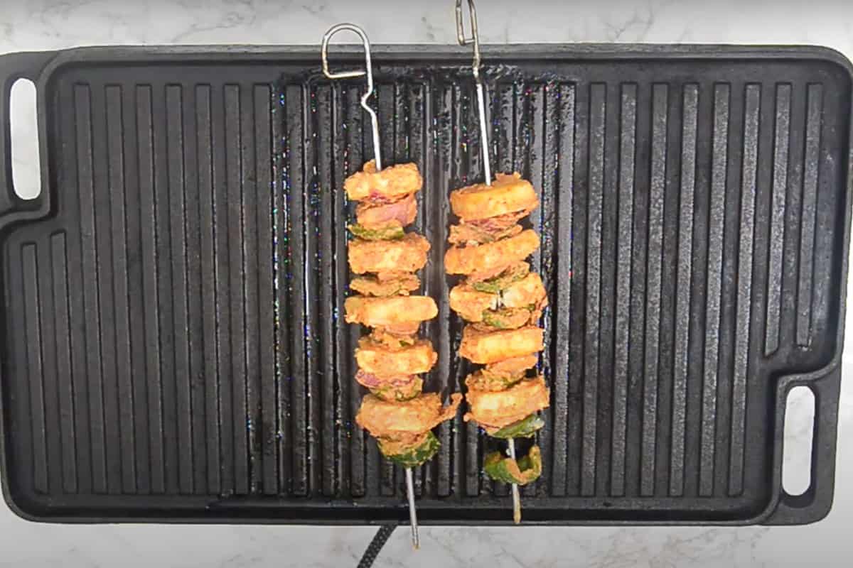 Paneer, capsicum and onion threaded on metal skewers and grilling on a grill pan.