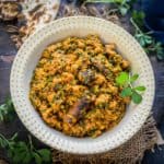 A white bowl filled with vibrant Methi Keema curry, garnished with spices and herbs, sits on a rustic surface alongside pieces of flatbread and fresh green leaves.