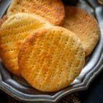 Four round, golden-brown Bakarkhani flatbreads are arranged on a silver platter, accompanied by a small bowl of ghee with a spoon. A piece of bread and blue cloth are visible in the background.