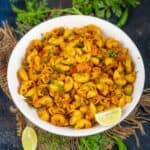 A white bowl filled with Masala Macaroni garnished with chopped herbs, surrounded by lemon wedges, green chilies, coriander leaves, and a red chili on a dark surface.