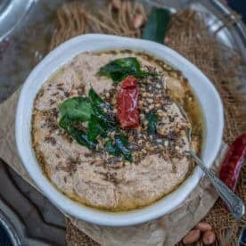 A white bowl of Andhra Style Peanut Chutney, garnished with fried curry leaves, dried red chili, and mustard seeds, sits on a rustic tray surrounded by scattered peanuts and spices.