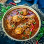 A bowl of spicy Chicken Angara curry garnished with fresh herbs and red chilies, surrounded by pieces of flatbread and a dark blue cloth on a rustic surface.