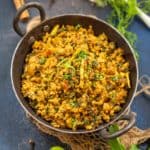 A black bowl filled with spicy Chicken Keema Masala curry, garnished with fresh coriander, placed on a rustic fabric mat. Green chili, mint leaves, spring onions, and whole spices are scattered around the bowl on a dark background.