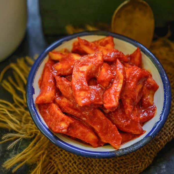 A white bowl with a blue rim filled with Hing ka Achar, placed on a piece of burlap fabric with a spoon in the background.