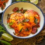 A white bowl with two pieces of spicy, golden-fried Egg Idli garnished with curry leaves and red chilies, surrounded by fresh cilantro, lime wedges, and a green chili on a rustic surface.