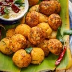 A plate lined with banana leaf holds several golden-brown Rava Appe with a bowl of creamy chutney garnished with red chilies and curry leaves, surrounded by whole dried red chilies.