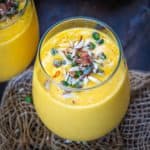 A close-up of Mango Lassi in two glasses, filled with creamy yellow goodness and topped with chopped nuts, saffron strands, and pistachios, set on a rustic burlap mat against a dark background.