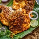 Three pieces of Pomfret Fry, spiced and grilled to perfection, are served on a banana leaf with sliced onions, lime, and fresh herbs on a rustic tray. The fish is coated in a rich reddish-brown spice mix.