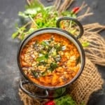 A bowl of Onion Tomato Chutney garnished with curry leaves, mustard seeds, and spices, placed on a burlap cloth with fresh cilantro and red chilies in the background.