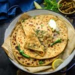 A plate of Broccoli Paratha topped with a dollop of butter, garnished with green chilies and cilantro, served with lemon wedges and a bowl of pickle on the side. A blue cloth is in the background.