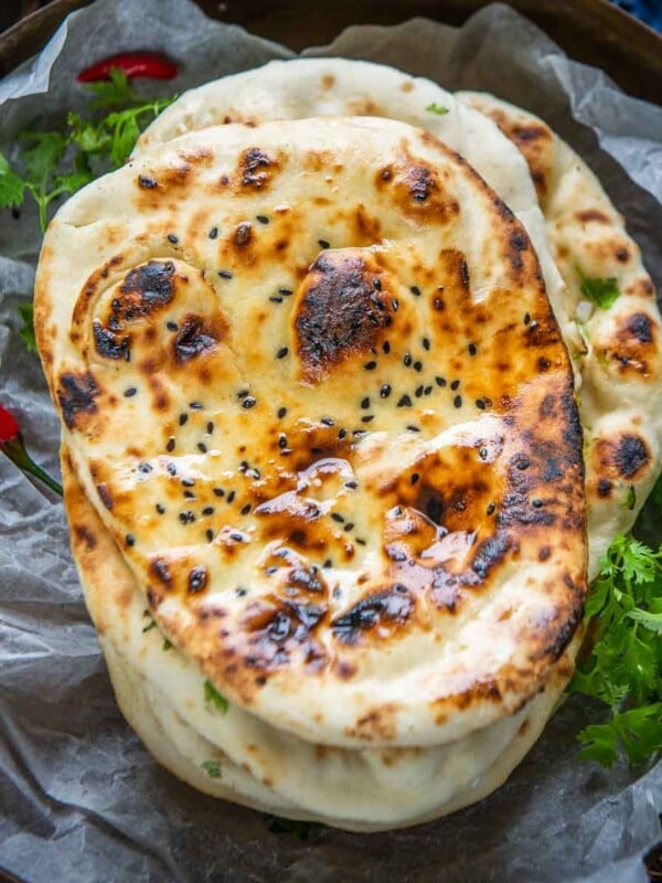 A close-up of a plate with three pieces of freshly baked butter naan, garnished with herbs and nigella seeds, placed on parchment paper with some green leafy garnish and red chili on the side.