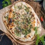 Two pieces of freshly baked Peshwari Naan topped with chopped herbs, nuts and seeds rest on parchment paper. Bowls with herbs, and whole chilies surround the bread on a dark surface.