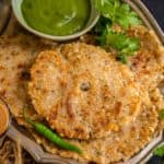 A plate of crispy Sabudana Thalipeeth garnished with cilantro, served with green chutney and two glasses of chai, sits on a dark surface with fresh coriander and a green chili.