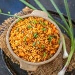 A bowl of Schezwan Fried Rice garnished with chopped green onions sits on burlap, with fresh spring onions beside it, all set on a dark rustic surface.
