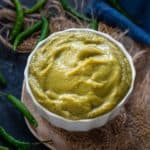 A white bowl filled with smooth Green Chilli Sauce sits on a piece of burlap, surrounded by whole green chilies on a dark surface, with a blue cloth in the background.