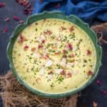 A bowl of creamy yellow sabudana kheer topped with sliced almonds, chopped pistachios, and rose petals is placed on a dark surface with some decoration around.