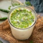 A glass of Taak (buttermilk) garnished with chopped coriander and spices sits on a textured mat, with green chilies and fresh coriander scattered nearby.