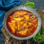 A bowl of Paneer Ghee Roast curry with triangular paneer pieces in a rich, red, spicy gravy, garnished with fresh herbs, on a rustic surface with green chilies and cilantro nearby.