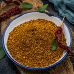 A bowl filled with coarse, golden-brown Karivepaku Podi sits on burlap, surrounded by dried red chilies and green curry leaves, with a blue cloth in the background.
