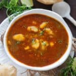 A white bowl of spicy aloo ki launji garnished with herbs, served alongside puffed puris, fresh coriander, a lime wedge, and a white spoon on a dark surface.