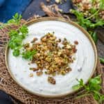 A bowl of creamy white Dried Nuts Raita topped with chopped nuts and herbs, garnished with fresh cilantro, placed on a rustic cloth with a dish of assorted nuts in the background.