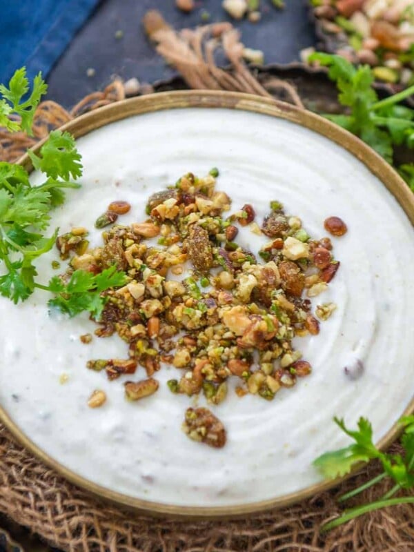 A bowl of creamy white Dried Nuts Raita topped with chopped nuts and herbs, garnished with fresh cilantro, placed on a rustic cloth with a dish of assorted nuts in the background.