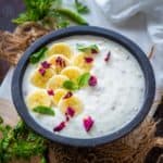 A bowl of creamy Banana Raita garnished with banana slices, fresh mint leaves, and rose petals, placed on a rustic surface with herbs and a green chili nearby.
