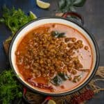 A bowl of spicy Kadala Curry garnished with curry leaves and dried red chili, surrounded by fresh herbs and lemon wedges on a rustic dark background.