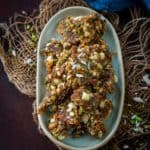 A plate filled with pieces of masala gur, a brittle snack made with mixed nuts, seeds, and jaggery, set on a rustic brown surface with jute fabric accents.