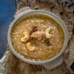 A bowl of brown Sakkarai Pongal garnished with cashew nuts and raisins, placed on a rustic mat with cardamom pods and cloves scattered around.