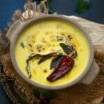 A bowl of yellow Mor Kuzhambu curry, topped with dried red chilies and black mustard seeds, garnished with fresh curry leaves, set on a rustic brown mat against a dark background.