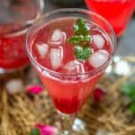 A glass of bright red Rose Soda iced drink garnished with a sprig of mint, with two similar drinks and scattered rose petals and mint leaves in the background.
