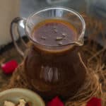 A glass pitcher filled with brown Thandai Syrup sits on a rustic tray, surrounded by rose petals. Nearby, a small bowl holds sliced almonds and pistachios, with a glass of creamy thandai garnished with petals in the background.