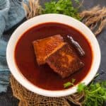 A white bowl of Kashmiri Lal Paneer curry, featuring two rectangular pieces of fried paneer and a whole dried chili, garnished with fresh cilantro, is placed on a rustic jute mat.