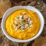 A white bowl filled with thick, bright yellow mango shrikhand, reminiscent of sweet aamras, is garnished with chopped nuts and sits on a rustic wooden board with pieces of crisp puri bread around it.