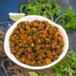 A white bowl filled with spicy Bhindi Bhaji stir-fry, garnished with herbs, sits on a burlap mat with lime wedges, green chili, cilantro, and lime in the background.