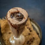 A tall glass of chocolate cold coffee topped with a scoop of chocolate ice cream and chocolate syrup, placed on a rustic tray with a brown cloth and chocolate chips in the background.