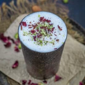 A tall metal glass of creamy lassi, topped with crushed pistachios and dried rose petals, rests on brown parchment paper scattered with nuts and petals.