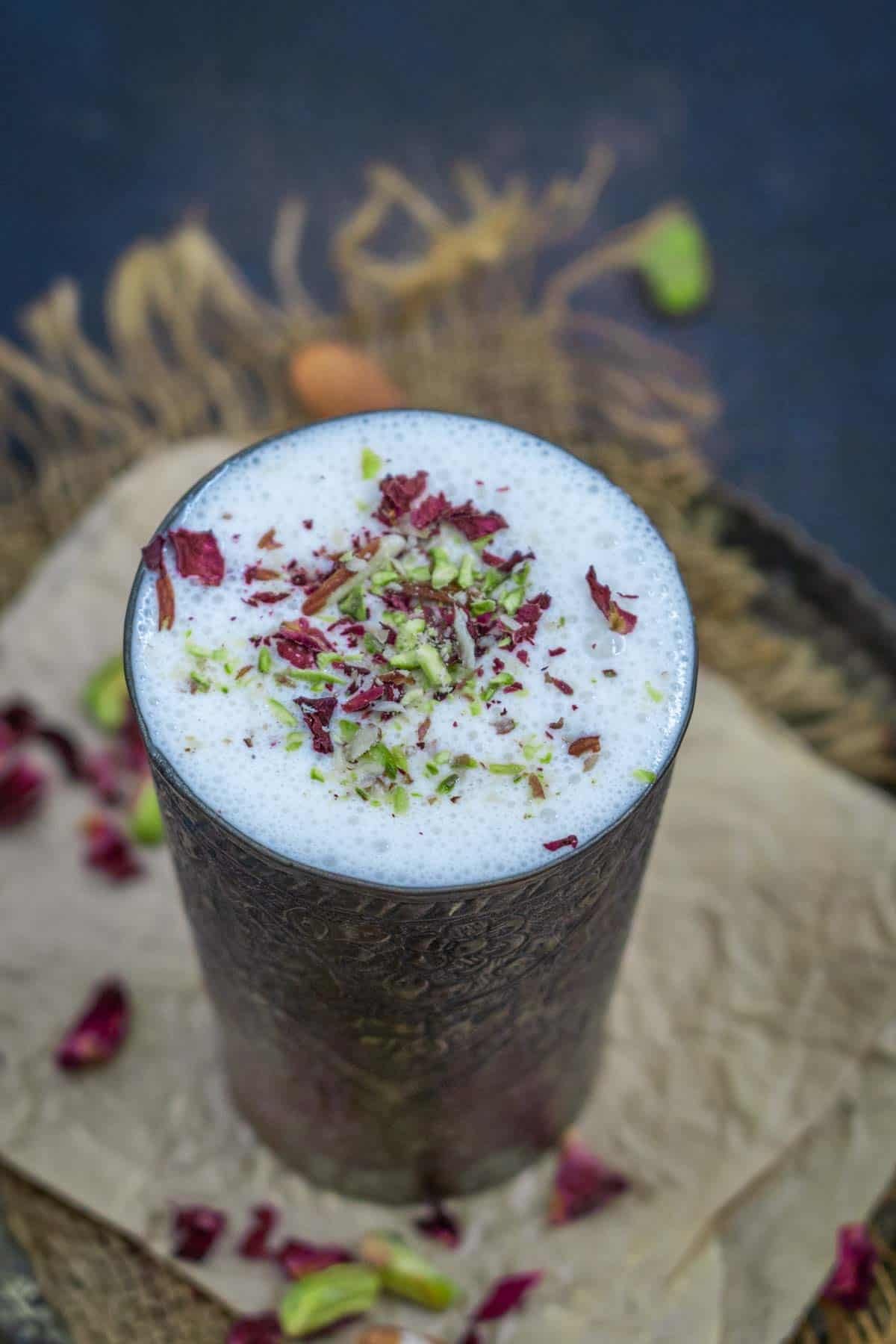 A tall metal glass of creamy lassi, topped with crushed pistachios and dried rose petals, rests on brown parchment paper scattered with nuts and petals.