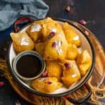 A white bowl filled with golden fried Kalakand Wontons, garnished with edible silver leaf and dried rose petals, served with a small container of dark dipping sauce on a wooden board.