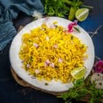 A white plate filled with yellow Poha Bhel, garnished with chopped red onions, cilantro, and lime wedges, sits on a dark surface surrounded by fresh herbs and a blue cloth.