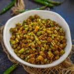 A white bowl filled with spicy Mirchi Ke Tipore, garnished with spices, sits on a rustic surface next to green chilies and burlap string.