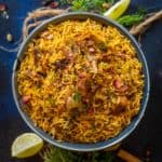 A bowl of flavorful Spicy Mutton Pulao garnished with herbs sits on a dark surface, surrounded by lemon wedges, spices, and fresh greens.